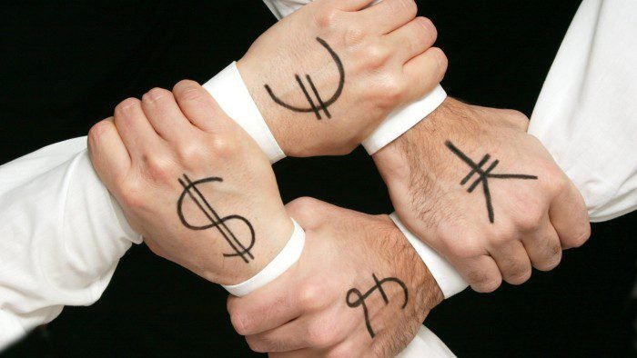 Four hands clasping each other’s write to form a square. The back of each hand has a currency symbol written on it - £ $ € and ¥