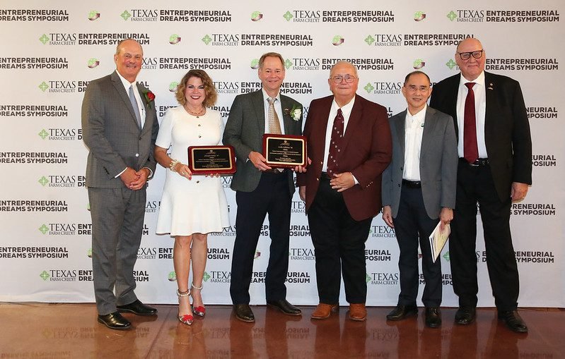 Six individuals standing, two of them holding awards, in from of a white backdrop that says Texas Farm Credit Entrepreneurial Dreams Symposium